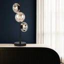 VARLIN A Modern table lamp with three silver glass spheres on a black base against a dark blue wall.